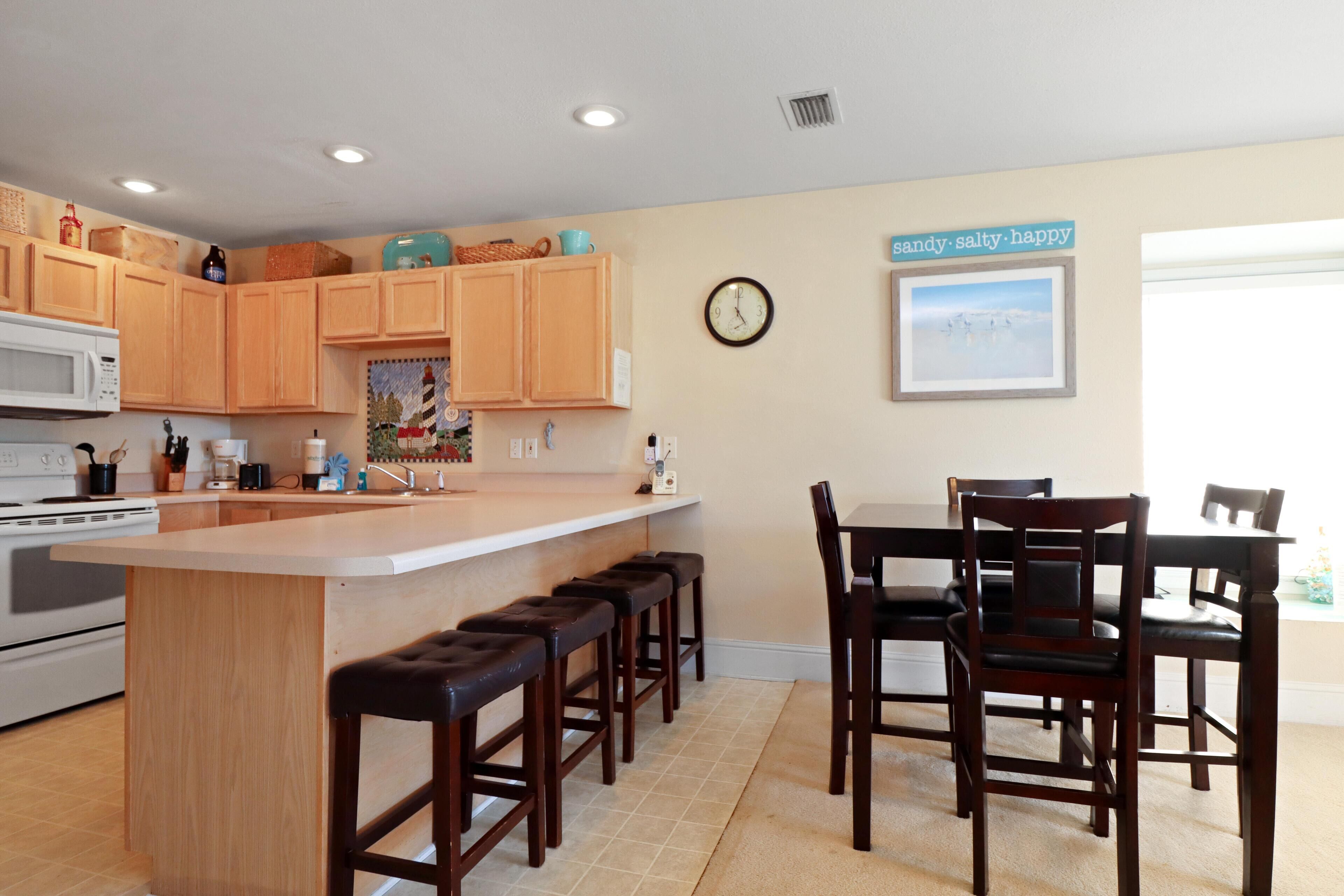 A kitchen with wooden cabinets, a central island with stools, a microwave, and a clock on the wall.