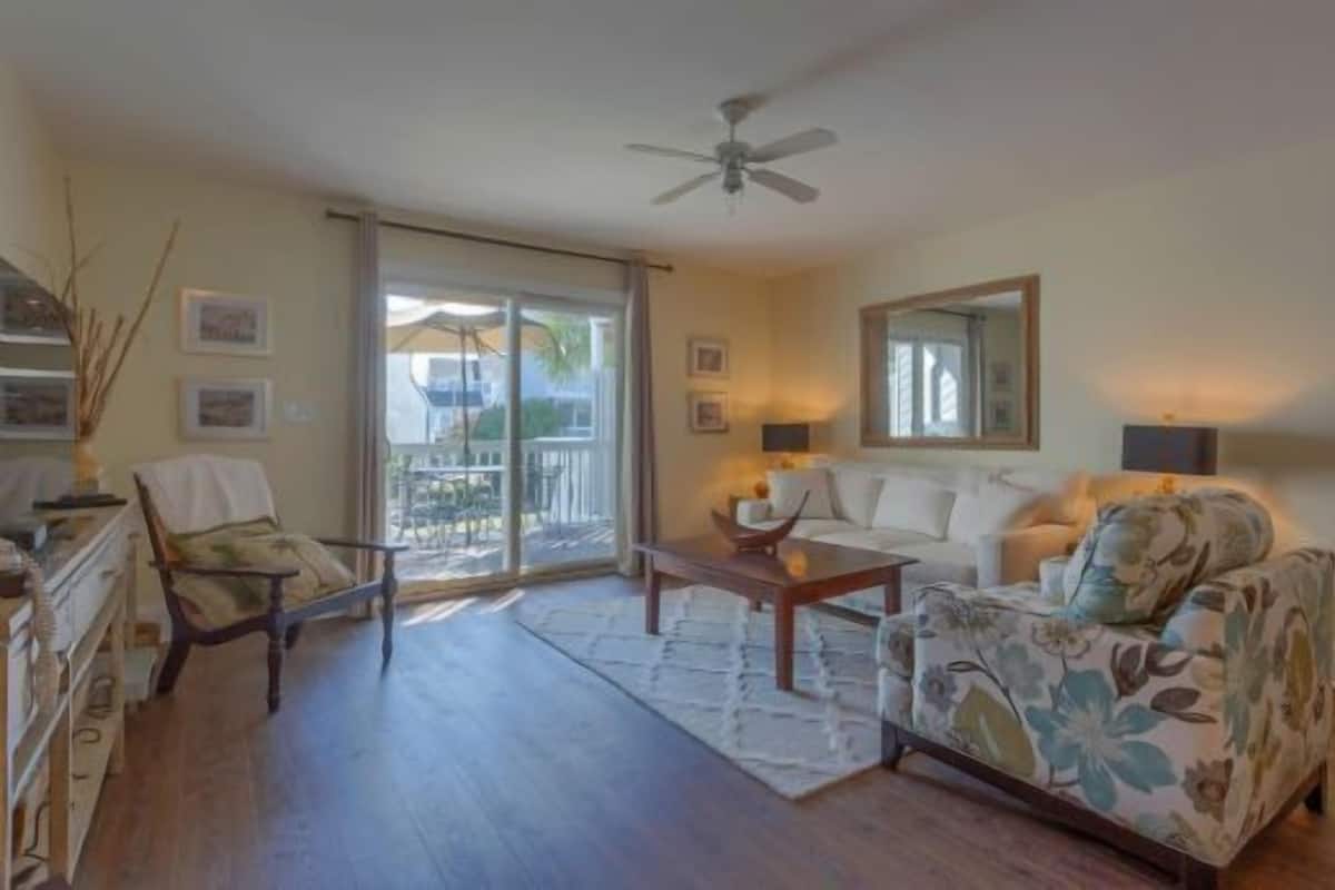 Sala de estar con sofá, sillón, mesa de centro y ventilador de techo. Hay una puerta corrediza de vidrio que da a un área exterior.