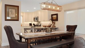 A dining area with a wooden table, dark chairs, and a bench. There is a kitchen in the background with white cabinets and a microwave. - Barrier Dunes 281 - 8 Branch Office (Port St. Joe)
