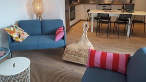 A modern living room with a blue sofa, a small round table, and a woven pouf. A kitchen with a dining area is visible in the background. - Gîte 10pers Avec Jacuzzi & Sauna Privatifs (Blainville-sur-Mer)