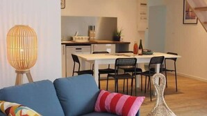 A modern living room with a blue sofa, a woven pouf, a small round table with books, and a kitchen in the background. - Gîte 10pers Avec Jacuzzi & Sauna Privatifs (Blainville-sur-Mer)