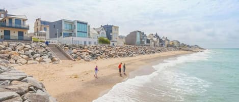 A beach with a rocky shoreline, a few people walking, and buildings along the coast.