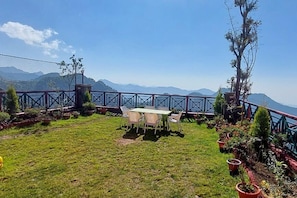 A grassy outdoor area with a table and chairs, surrounded by potted plants and a metal fence overlooking a mountainous landscape.