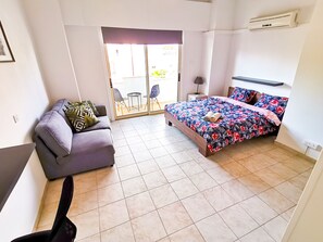 A bedroom with a bed, a sofa, a chair, a small table, and a sliding glass door leading to an outdoor area. - Central Square Nomadic Rooms (Larnaca)