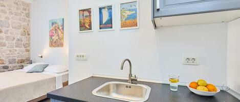Una cocina compacta con un lavamanos, un bol de fruta y una cama en la habitación contigua.
