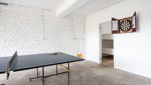 Una mesa de ping pong en una habitación con paredes de ladrillo blanco, un dardo y un reloj.