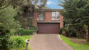 A two-story brick house with a garage, surrounded by greenery and a paved driveway.