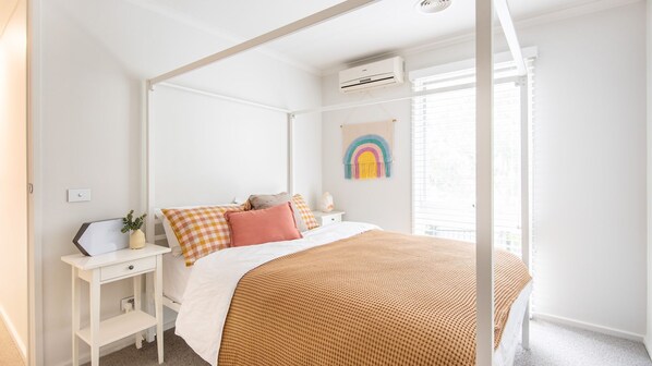 A bedroom with a four-poster bed, white walls, a small bedside table with a lamp, and a window with blinds.