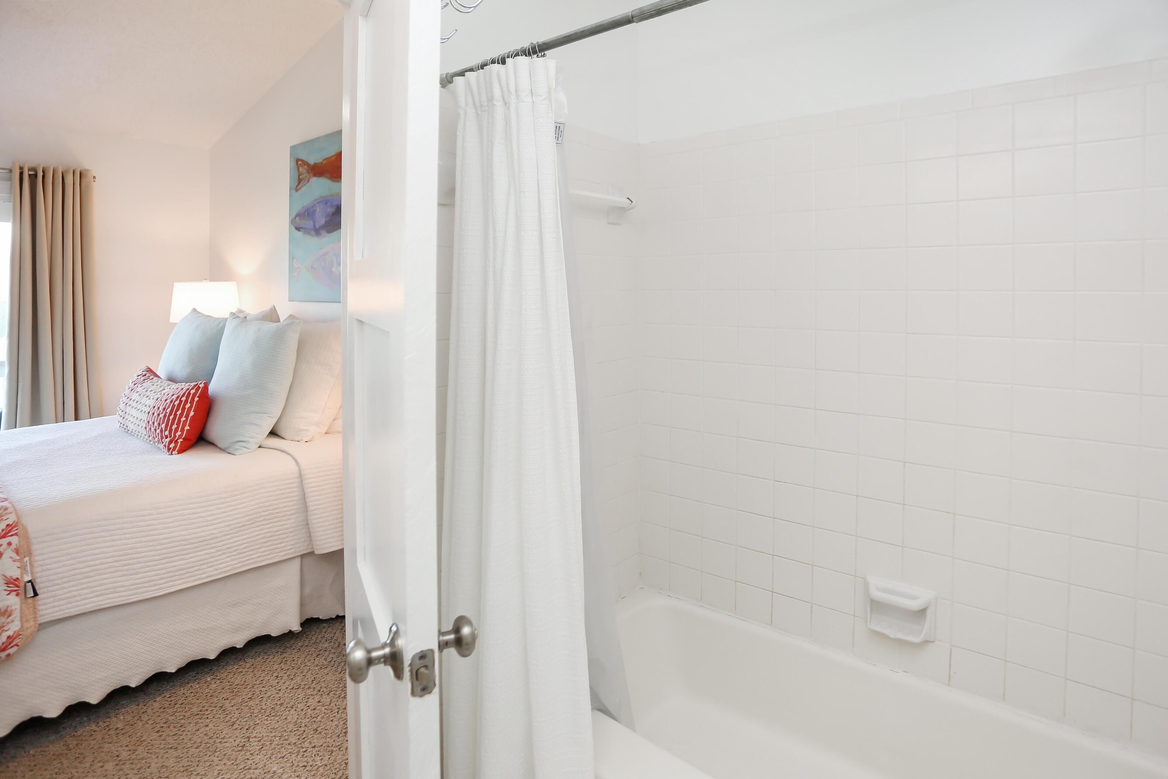 A bedroom with a bed, pillows, a nightstand, a bathroom with a bathtub, and a shower curtain.