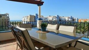 Un balcón con una mesa y sillas, una planta en maceta, y vistas a la ciudad y al mar.