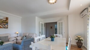 Un comedor con una mesa y sillas blancas, un sofá con cojines a rayas, una planta en maceta y un cuadro en la pared.