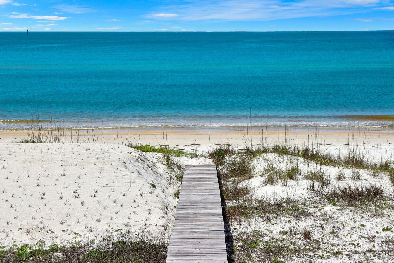 Une promenade en bois mène à une plage de sable fin bordée d’une eau d’un bleu limpide.
