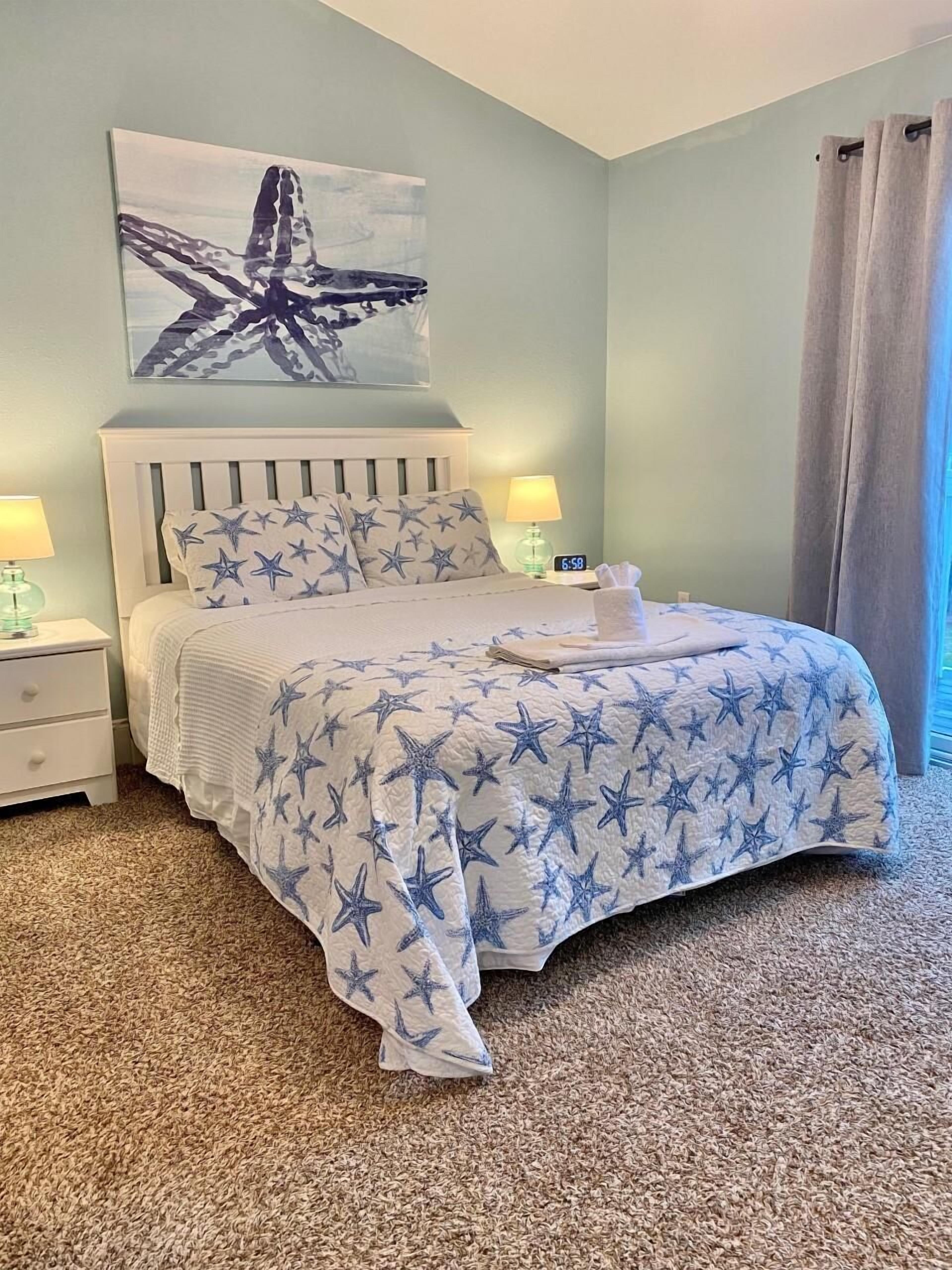 A bedroom with a bed, star-patterned bedding, a nightstand with a lamp, a wall clock, and a painting.