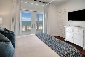 A bedroom with a bed, a dresser, a television, and a door leading to a balcony with a view of the sea.