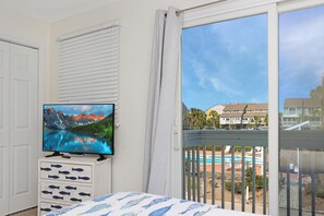 A bedroom with a TV on a dresser, a bed with a fish-patterned bedspread, a window with a view of buildings and a pool. - Barrier Dunes 248 - 111 Endless Summer (Port St. Joe)