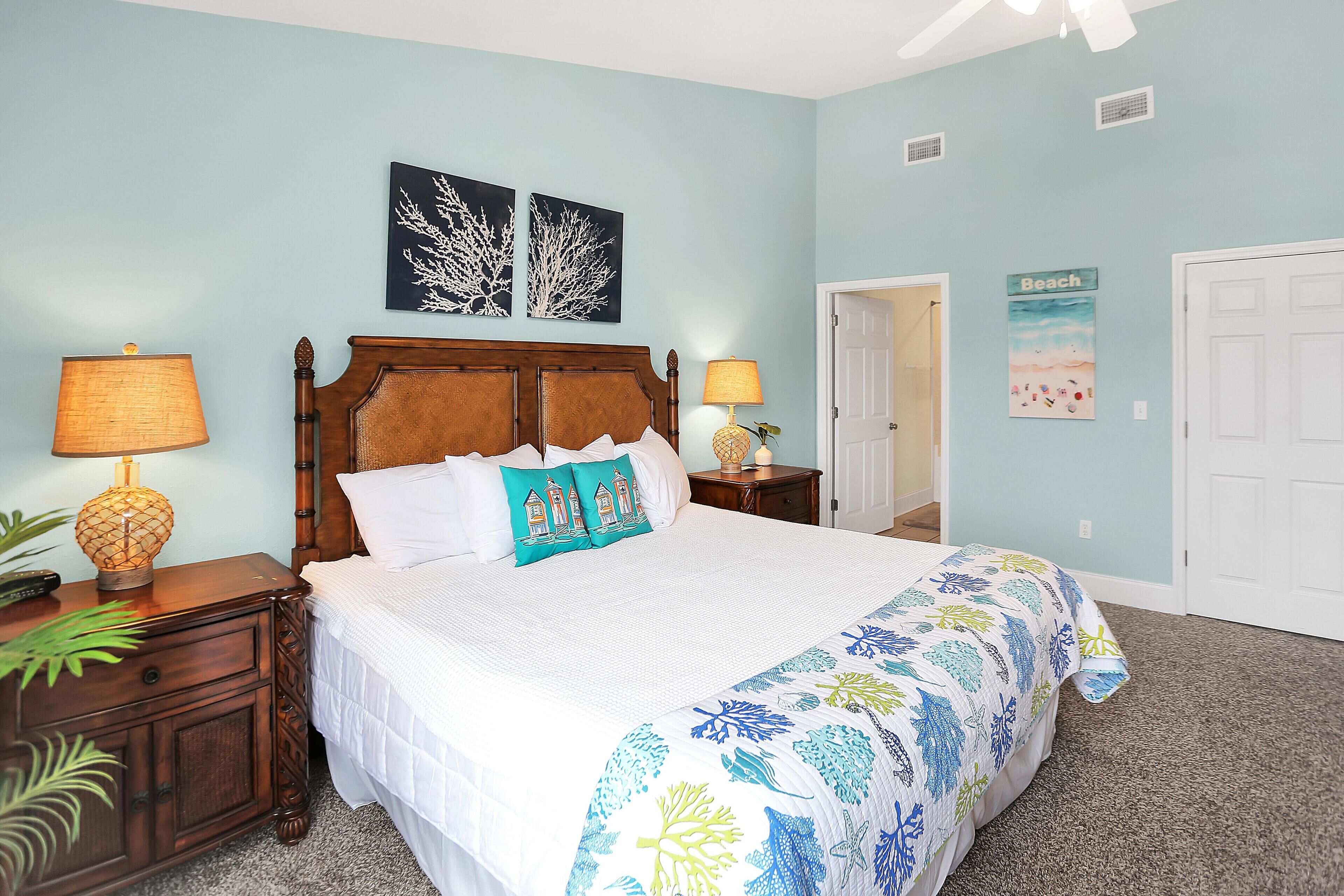 A neatly made bed with a white comforter and blue floral bedspread, a wooden nightstand with a lamp, and a wall with two framed pictures and a 'Beach' sign.