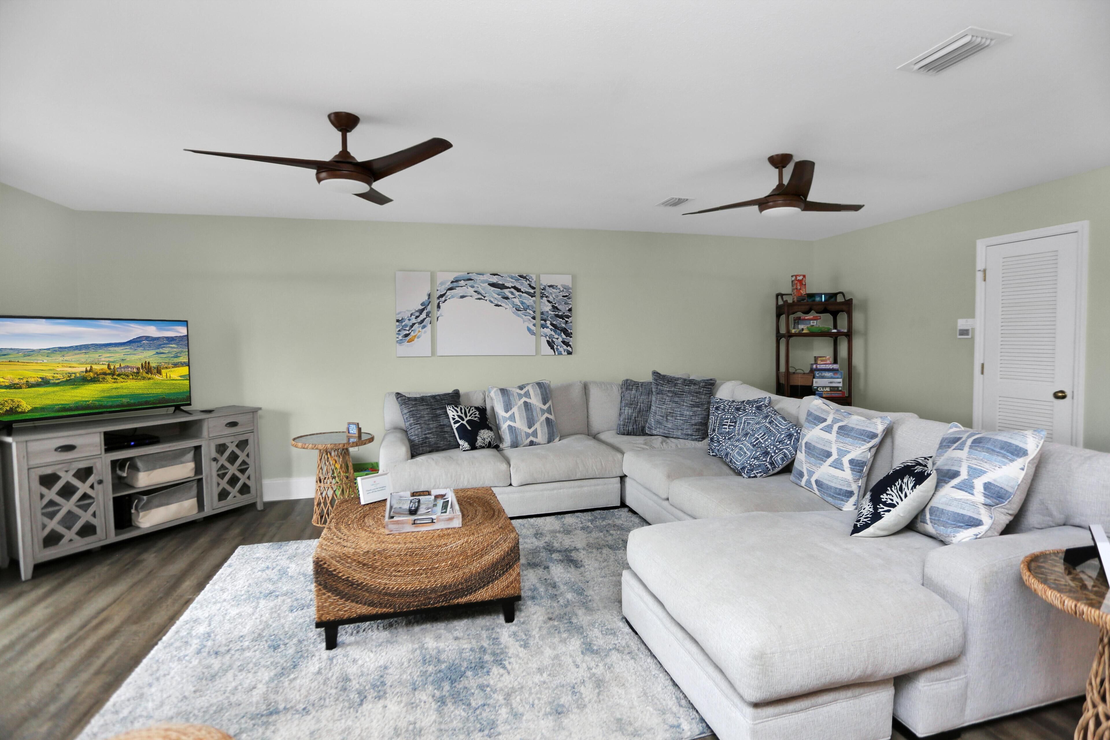 A living room with a sectional sofa, a flat-screen TV, ceiling fans, and a decorative wall piece.