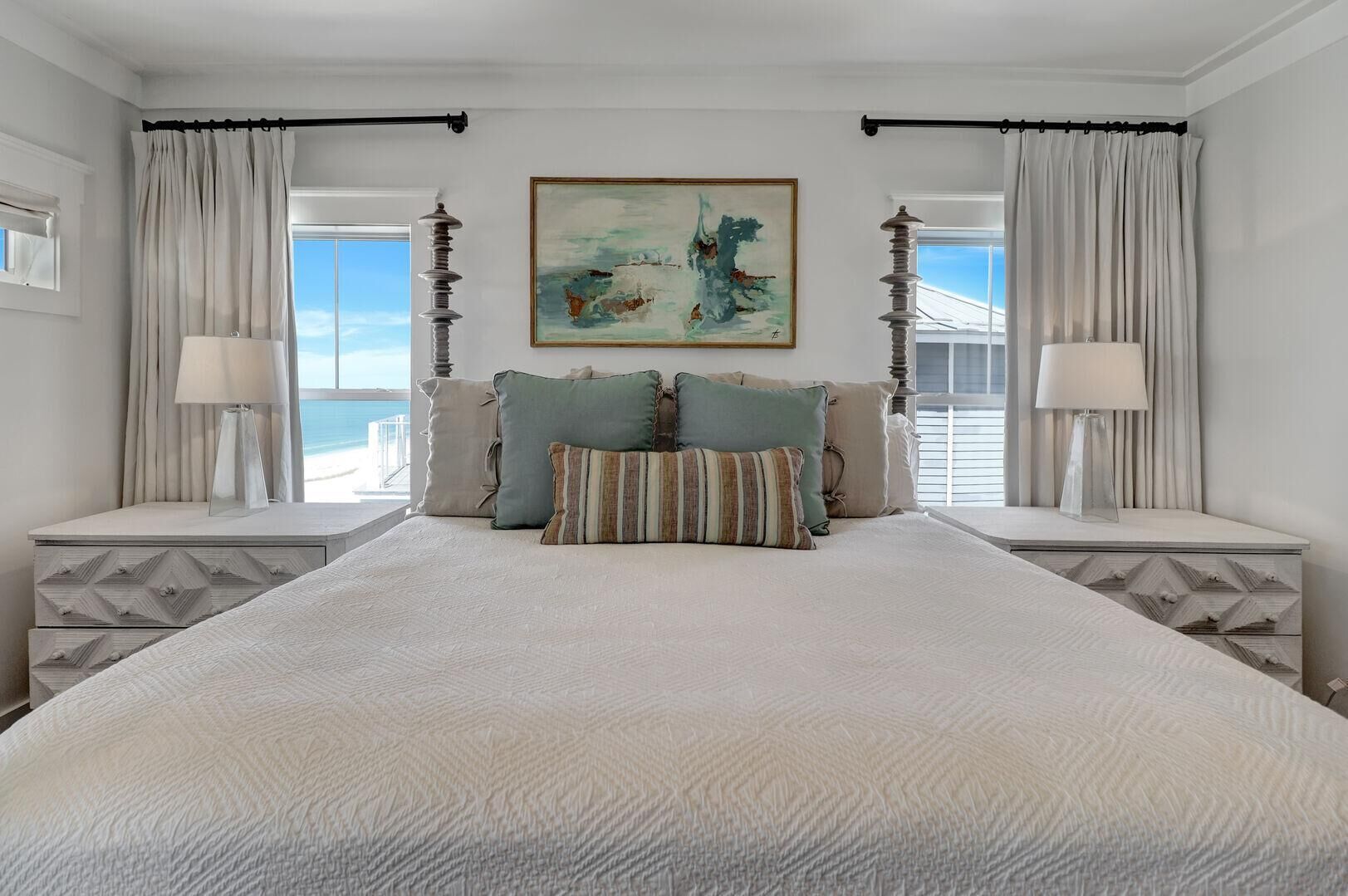 A bedroom with a large bed, two nightstands, a painting, and a view of the ocean.
