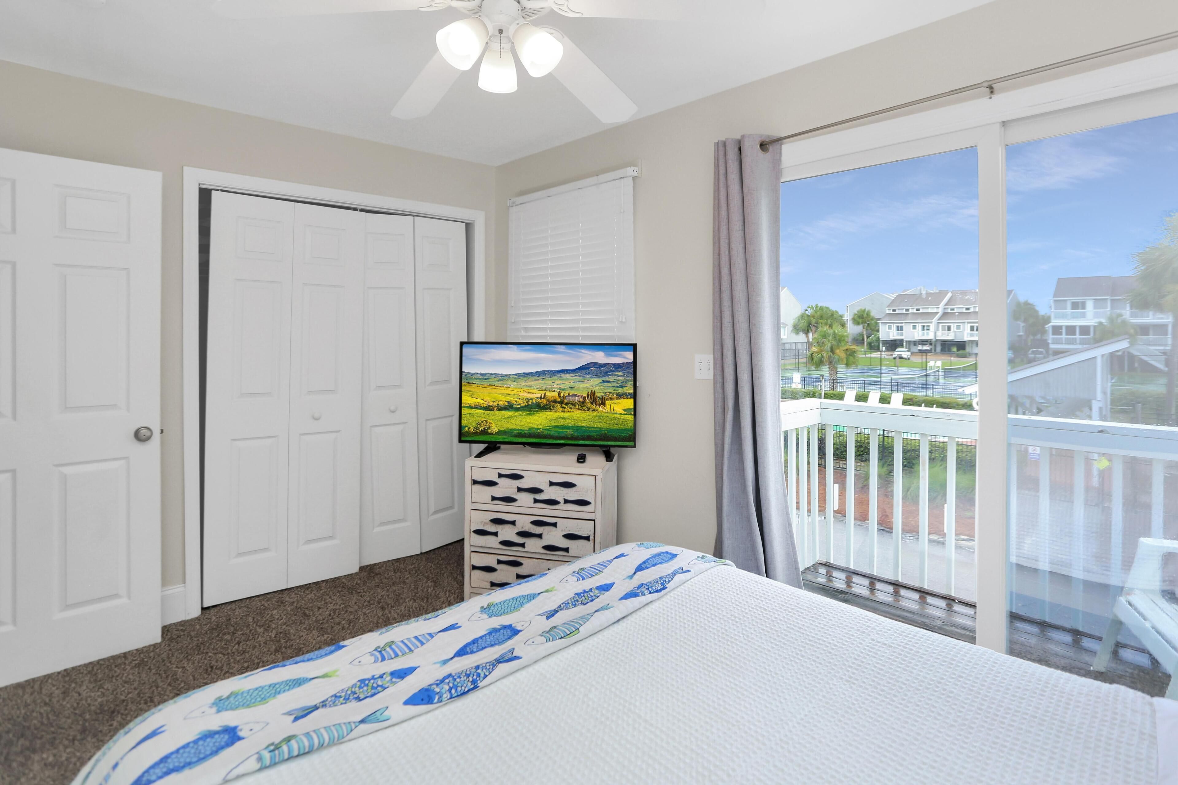A bedroom with a bed, a television, a ceiling fan, a sliding door, and a balcony with a view of houses.