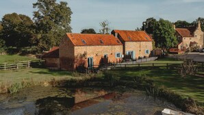 Un bâtiment traditionnel en pierre, surmonté d’un toit de tuiles rouges, entouré d’une végétation luxuriante et bordé d’un plan d’eau paisible.