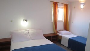 Une chambre avec deux lits, l’un recouvert d’une couvre-lit bleue et l’autre d’une couvre-lit blanche. Il y a une fenêtre avec des rideaux orange et un luminaire fixé au mur.