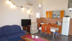 Un salon avec un canapé bleu, une table basse en verre et un téléviseur. Il y a également un coin cuisine avec des placards orange et un réfrigérateur.