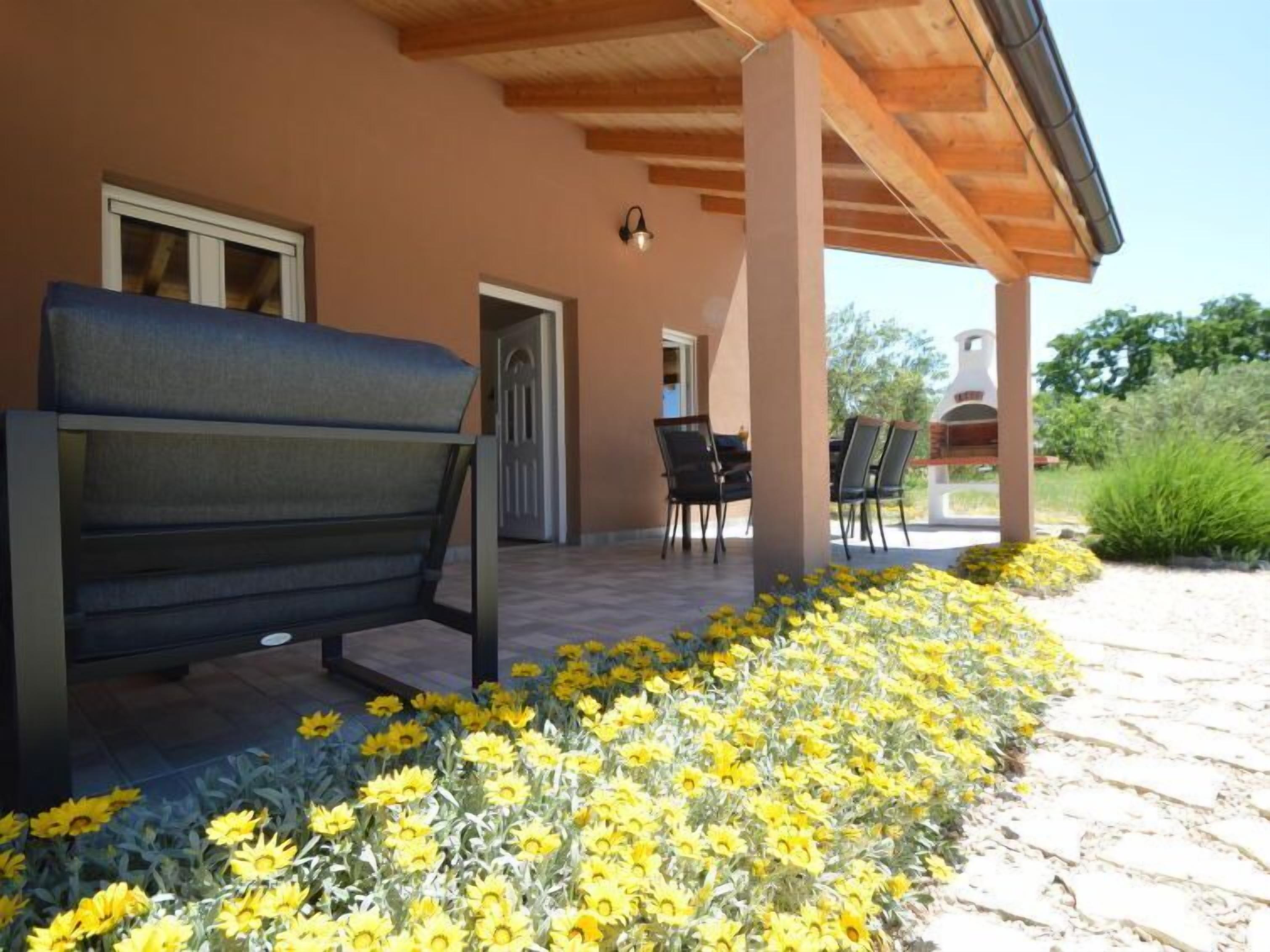 Un espacio al aire libre techado con un banco y sillas, rodeado de flores amarillas.