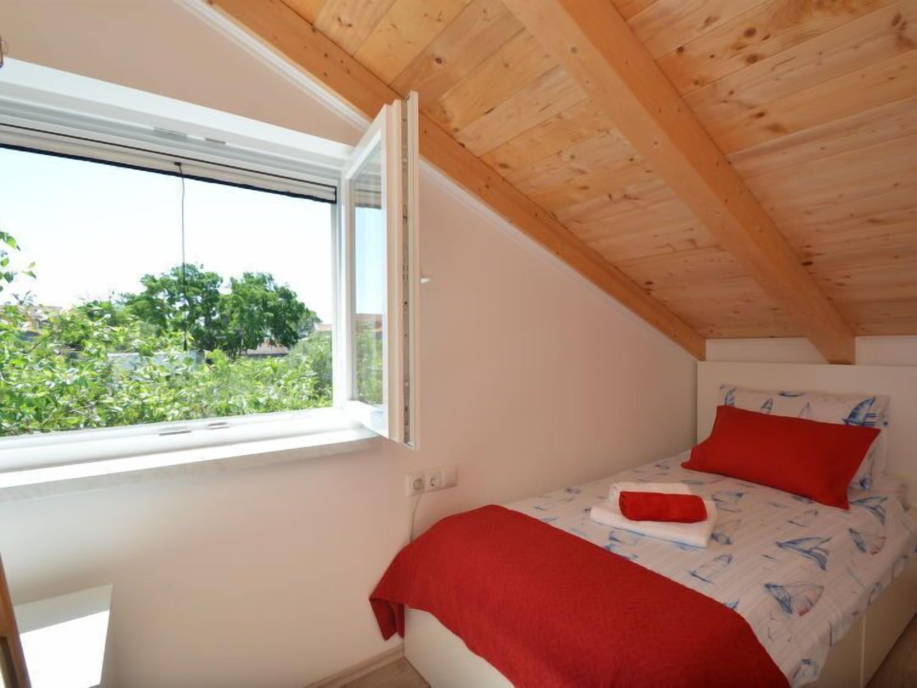 A small, single-bed room with a sloped ceiling and a window overlooking greenery.