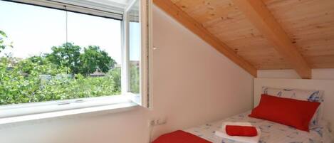 A small, single-bed room with a sloped ceiling and a window overlooking greenery.