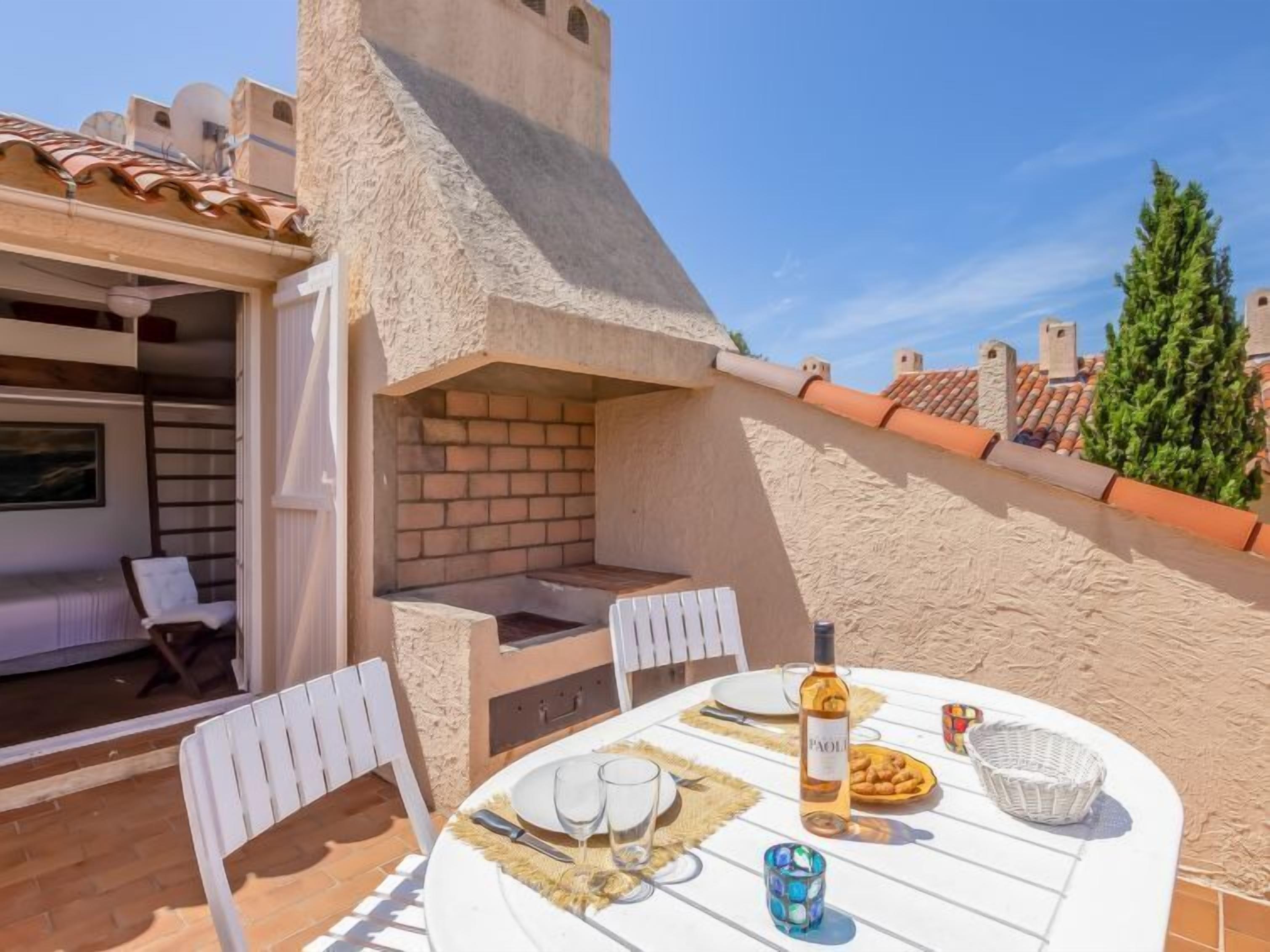 Une terrasse avec un four en briques, des chaises blanches, une table dressée pour le repas et une bouteille de vin.