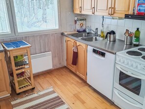 Une cuisine avec des meubles en bois, un lave-vaisselle, une cuisinière, une fenêtre avec des stores et un petit chariot en bois équipé d’un panneau solaire.
