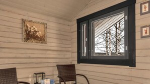 A room with a wooden paneled wall, a window with a black frame, two wicker chairs, a small table with a basket, and a framed picture on the wall. - Koivulahden reinola by Interhome (Ylitornio)