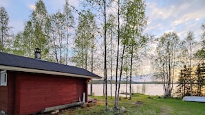 A red cabin with a small porch, surrounded by a grassy yard and birch trees, overlooking a calm lake. - Koivulahden reinola by Interhome (Ylitornio)