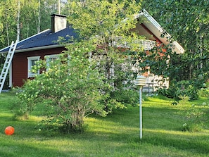 Une maison à l’extérieur rouge, avec un toit noir et une fenêtre blanche. Un arbre aux feuilles vertes se trouve devant la maison.
