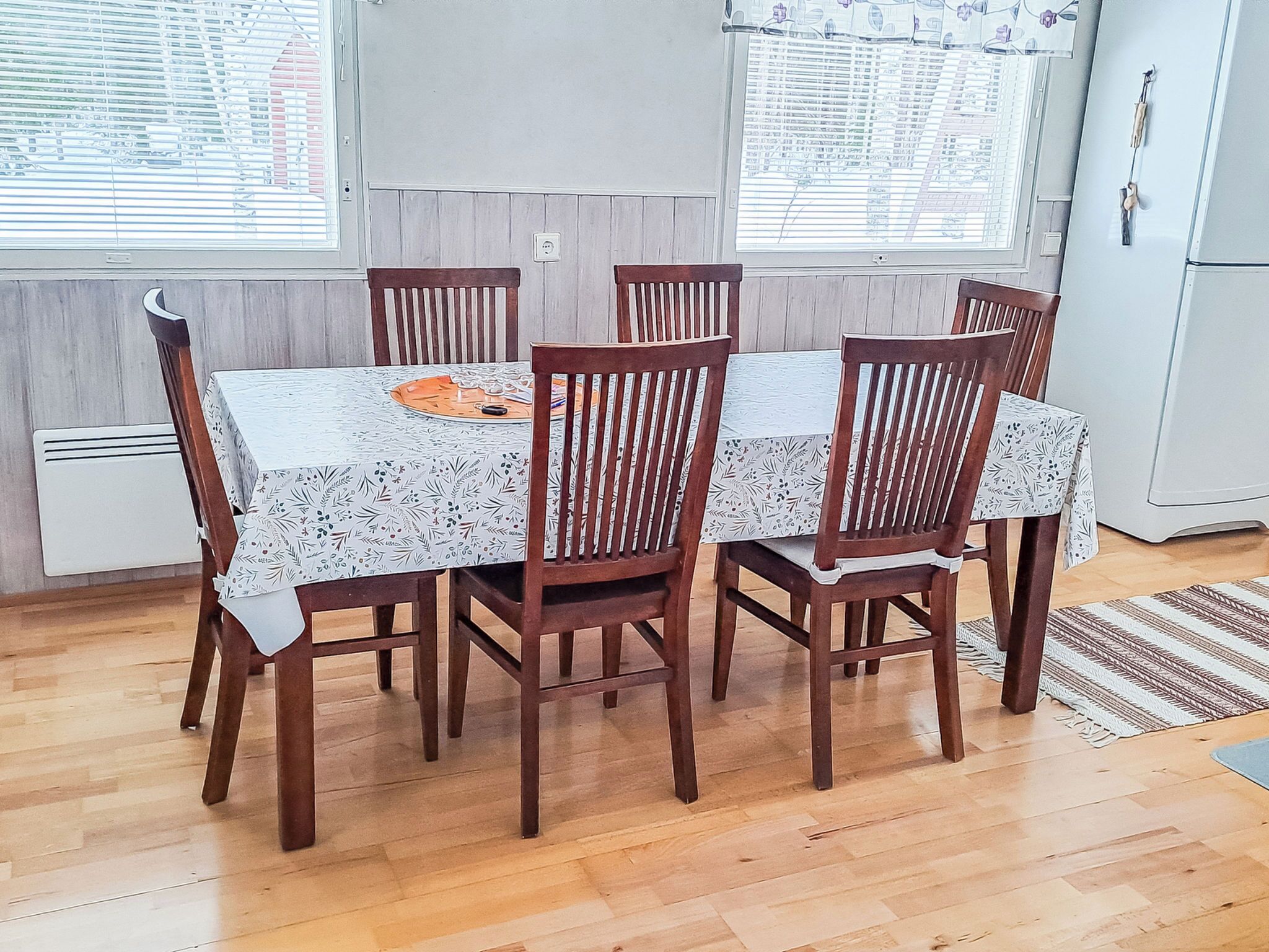A dining area with a table and six chairs, a window with blinds, a white radiator, and a refrigerator.