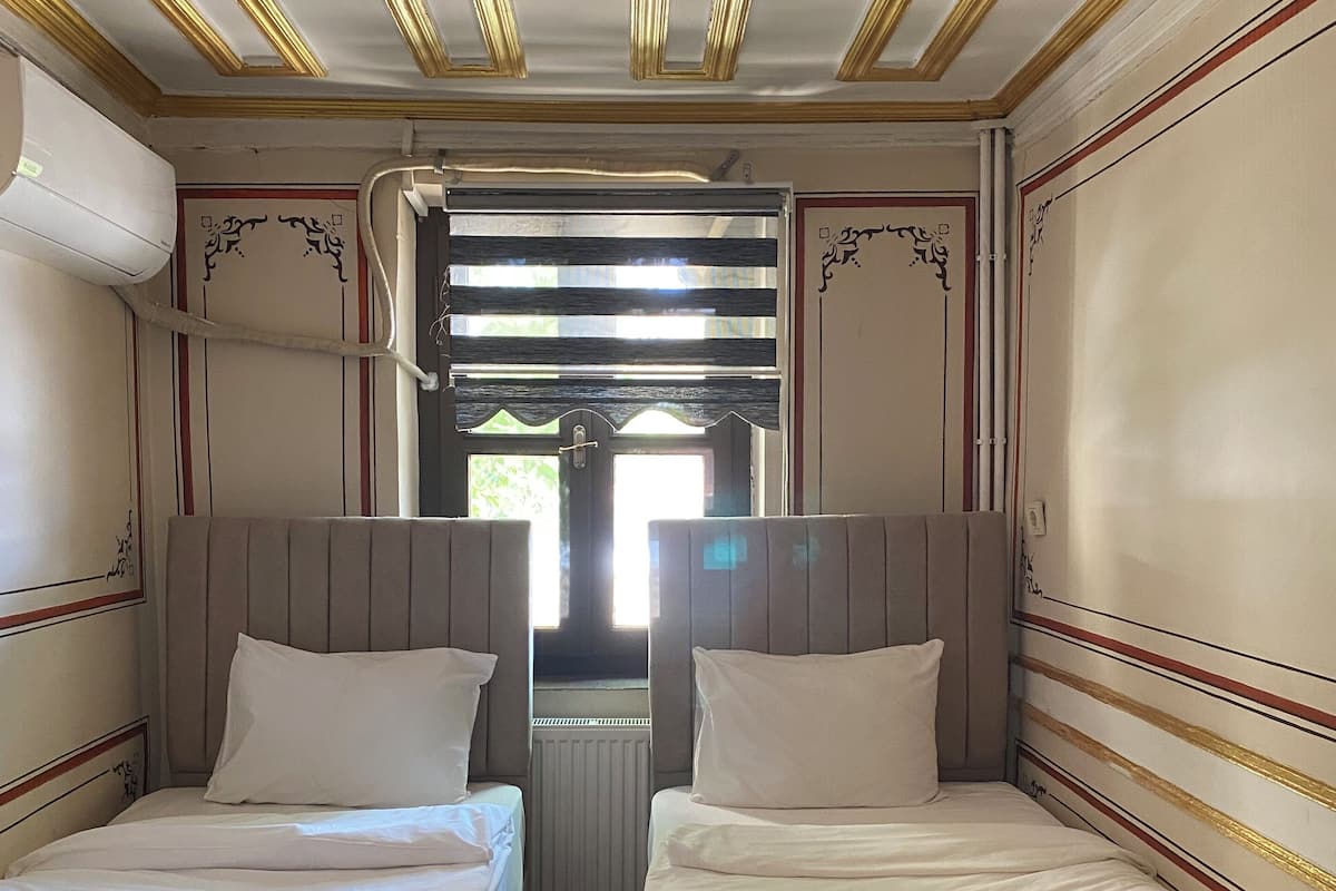 A hotel room with two beds, each with a pillow and a rolled towel. The room has a wooden floor and a radiator under the window.