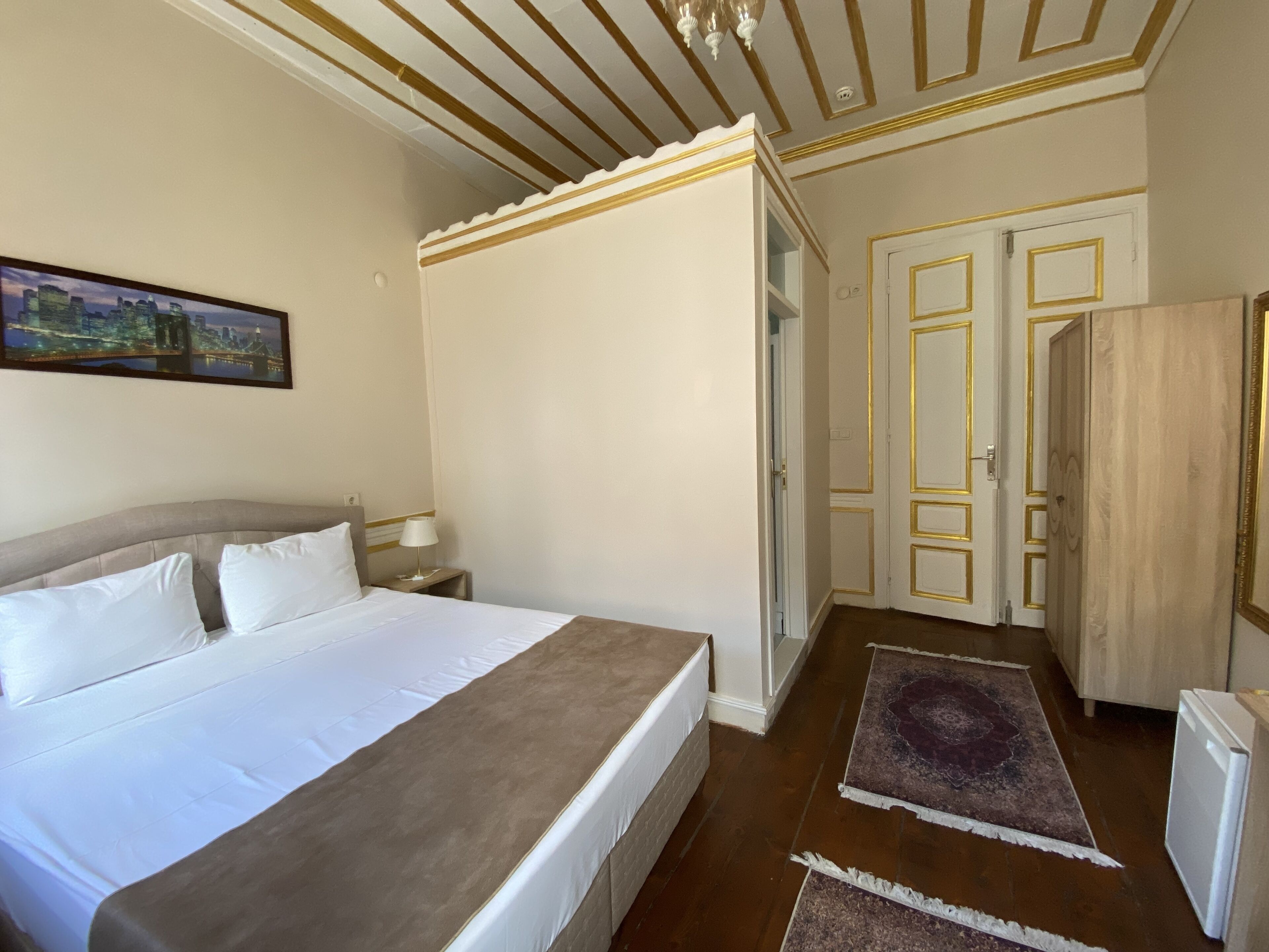 Habitación de hotel con una cama grande, una mesita de noche, una lámpara, un cuadro en la pared y una puerta que da a otra habitación.