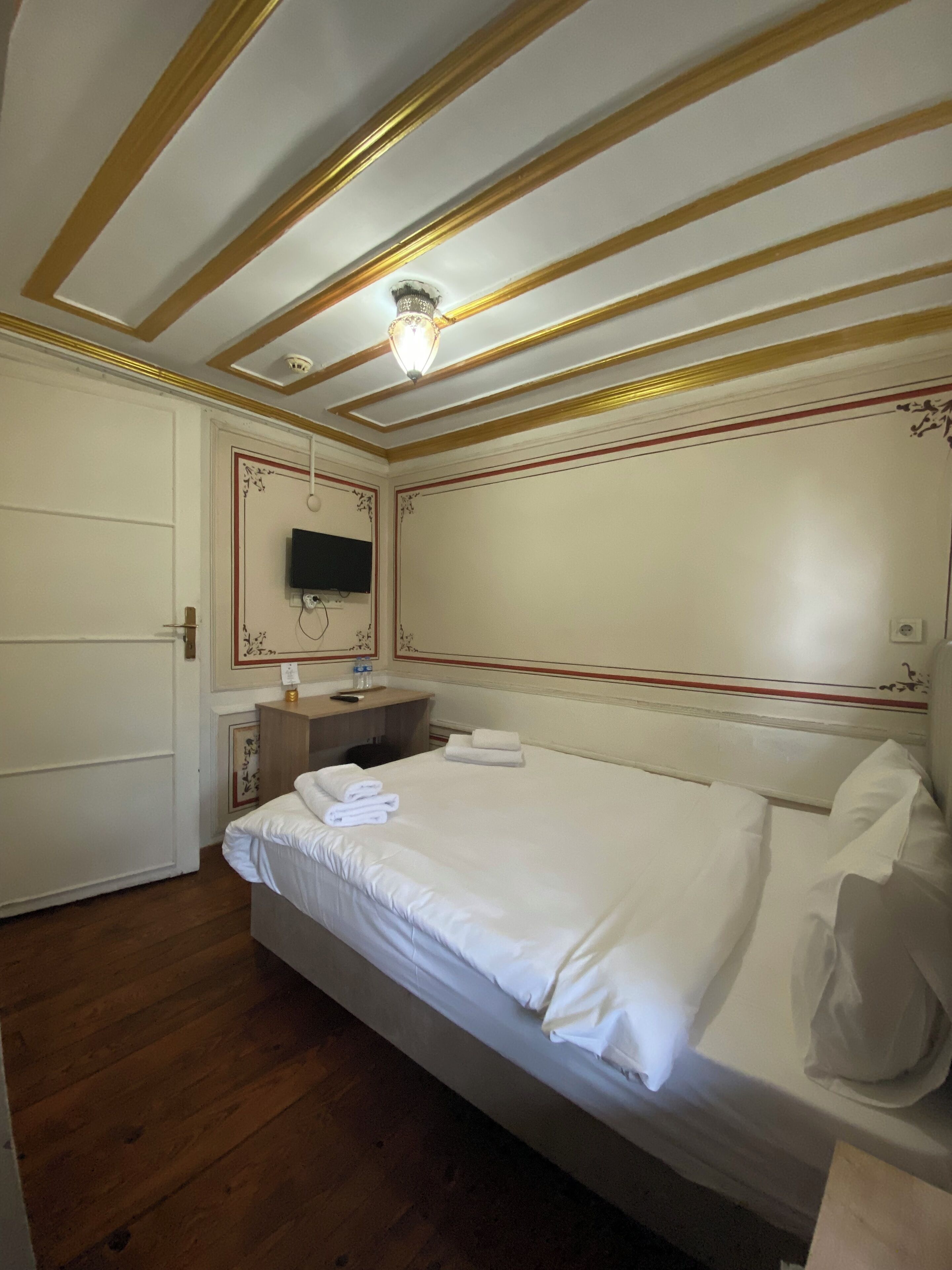 A hotel room with a bed, wooden floor, a desk, a chair, a television, and a ceiling light.