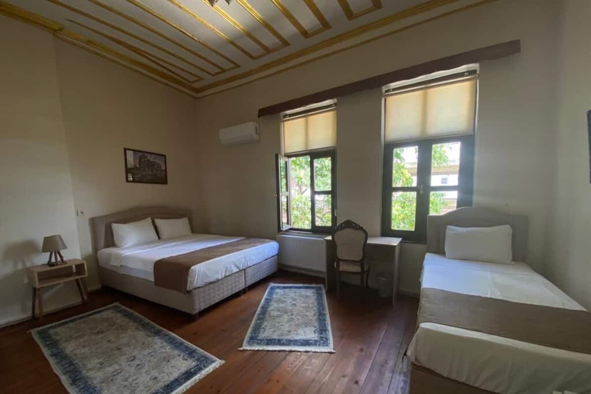 A room with two beds, a desk, and a chair. There are two windows providing natural light.