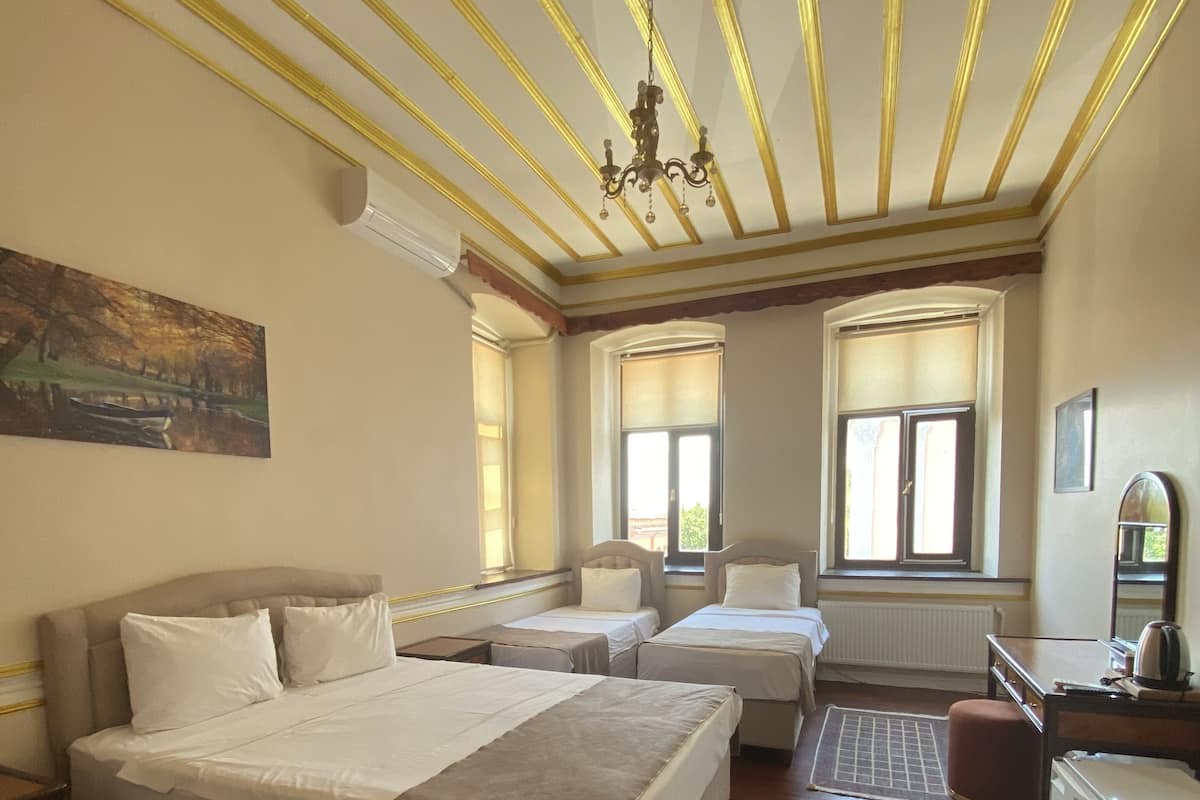 A bedroom with a large bed, two smaller beds, a desk, and a chair. There are windows with blinds, a radiator, and a chandelier.