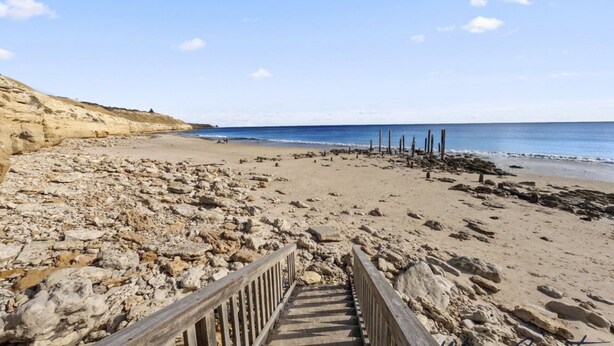 Ein Strand mit einer Holztreppe, die zum Ufer führt, felsigem Gelände und blauem Himmel.