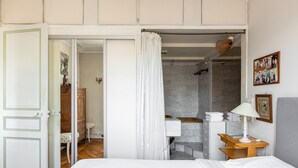 Ein Schlafzimmer mit Bett, einem hölzernen Nachttisch, einem Badezimmer mit Dusche und einer Tür zu einem anderen Raum.