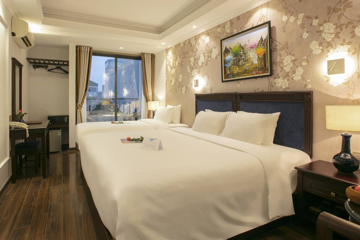 A hotel room with a large bed, a desk with a telephone, a chair, a small table with a lamp, and a window with curtains.