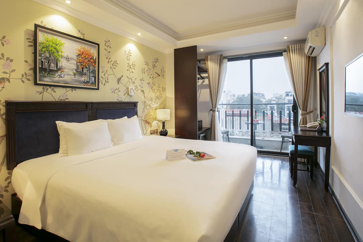 A hotel room with a large bed, a desk with a chair, a window with a view of the city, and floral wallpaper.
