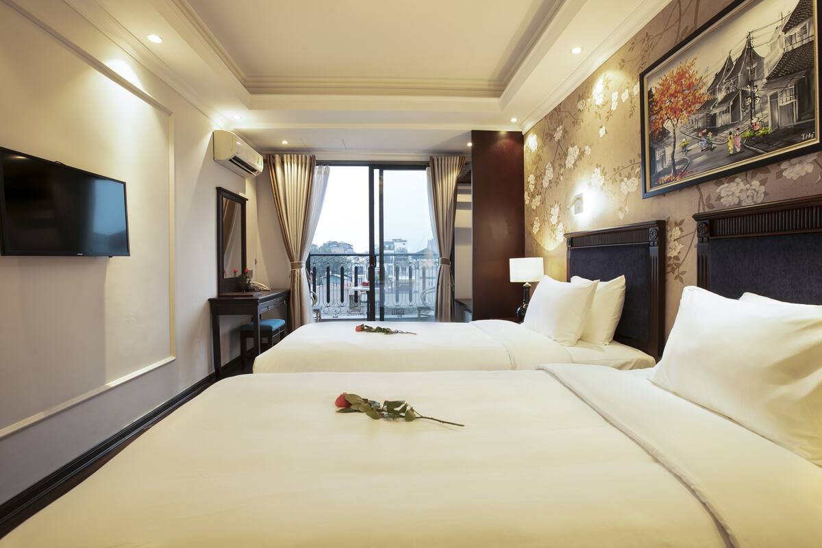 A hotel room with two beds, a TV, a balcony with a view, and a painting on the wall.