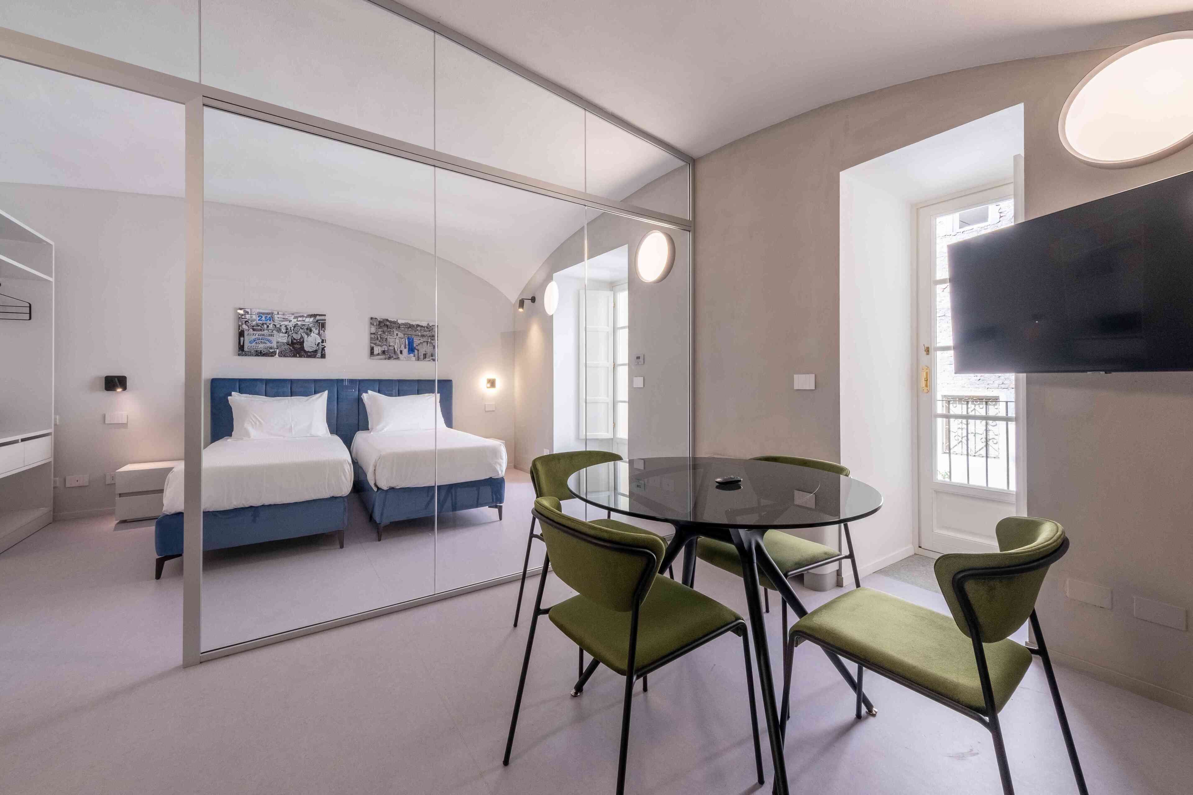 Habitación de hotel moderna con un separador de vidrio, una mesa de comedor redonda, sillas verdes, una cama con ropa de cama azul y un televisor montado.