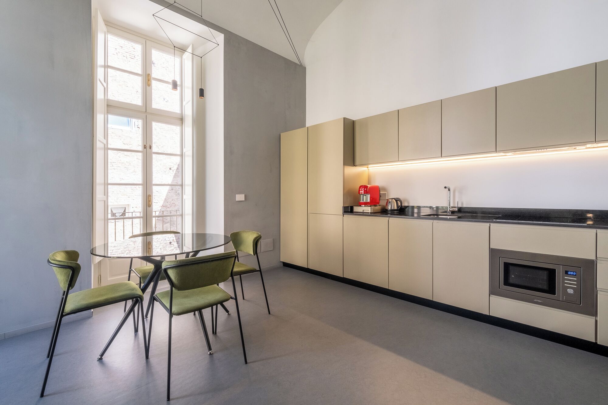 A modern kitchen with a round glass table and green chairs, a red coffee maker, and a built-in microwave.
