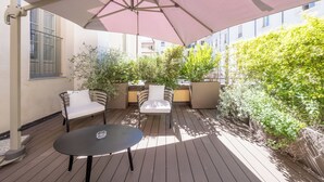 Un patio con una mesa y dos sillas bajo un paraguas grande, rodeado de plantas en macetas y una terraza de madera.