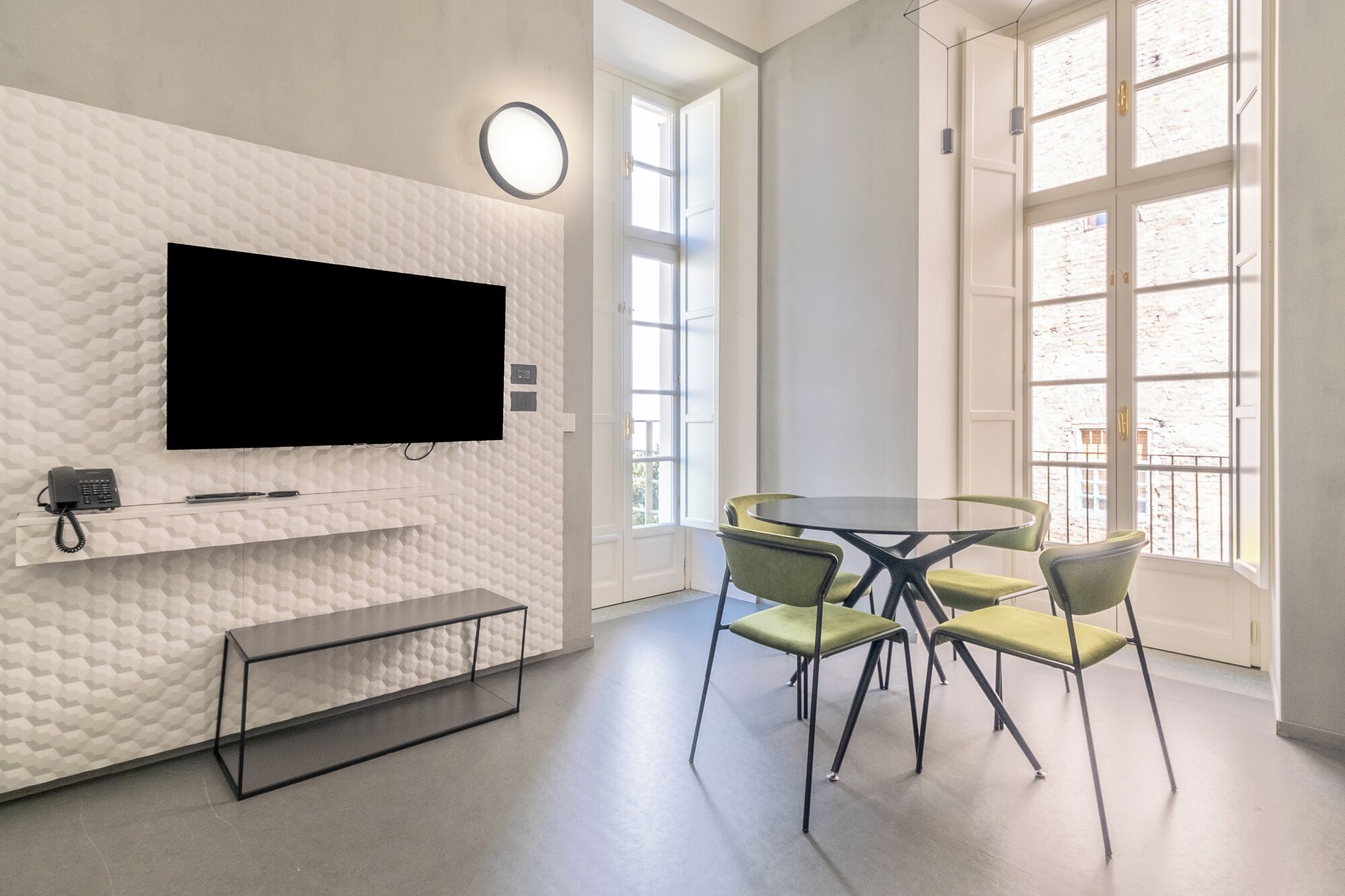 A modern room with a large flat-screen TV mounted on a textured wall, a small desk with a phone and pen, a round table with four chairs, and large windows with a view of buildings.