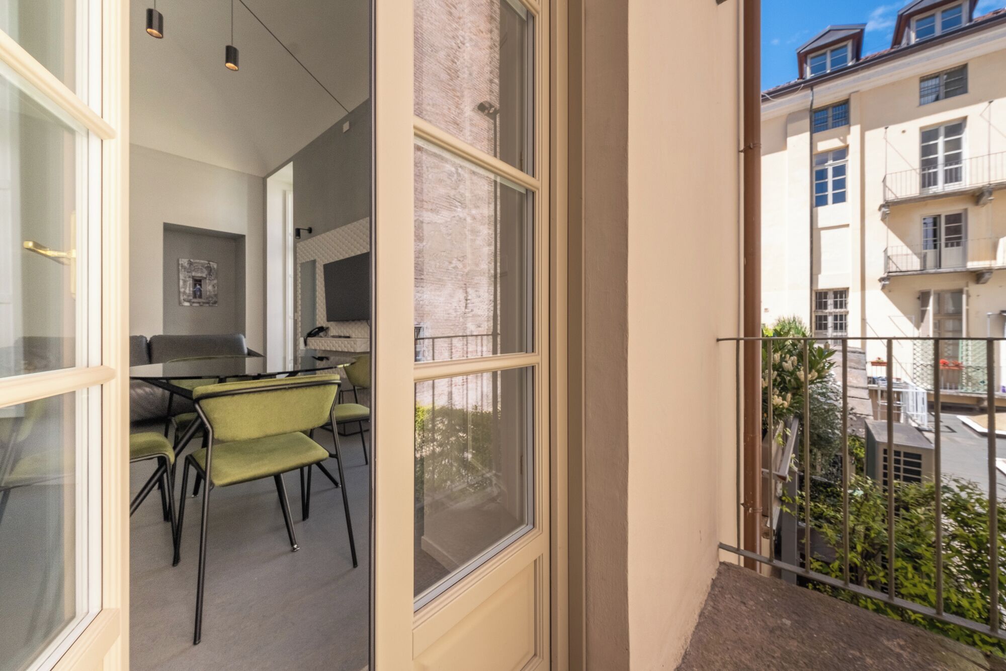A modern apartment with a balcony, a dining area with green chairs, and a kitchen visible through the window.